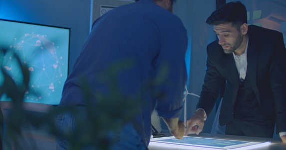 Two men in an office discuss a project displayed on an illuminated table, with a digital network graphic visible on a screen in the background.