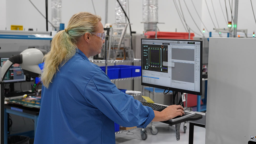 A technician wearing safety glasses and a blue uniform monitors diagnostic software on a computer at a workstation.
