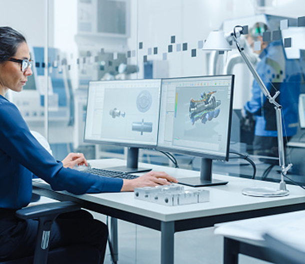 An engineer in an office uses dual monitors to work on 3D mechanical CAD designs.