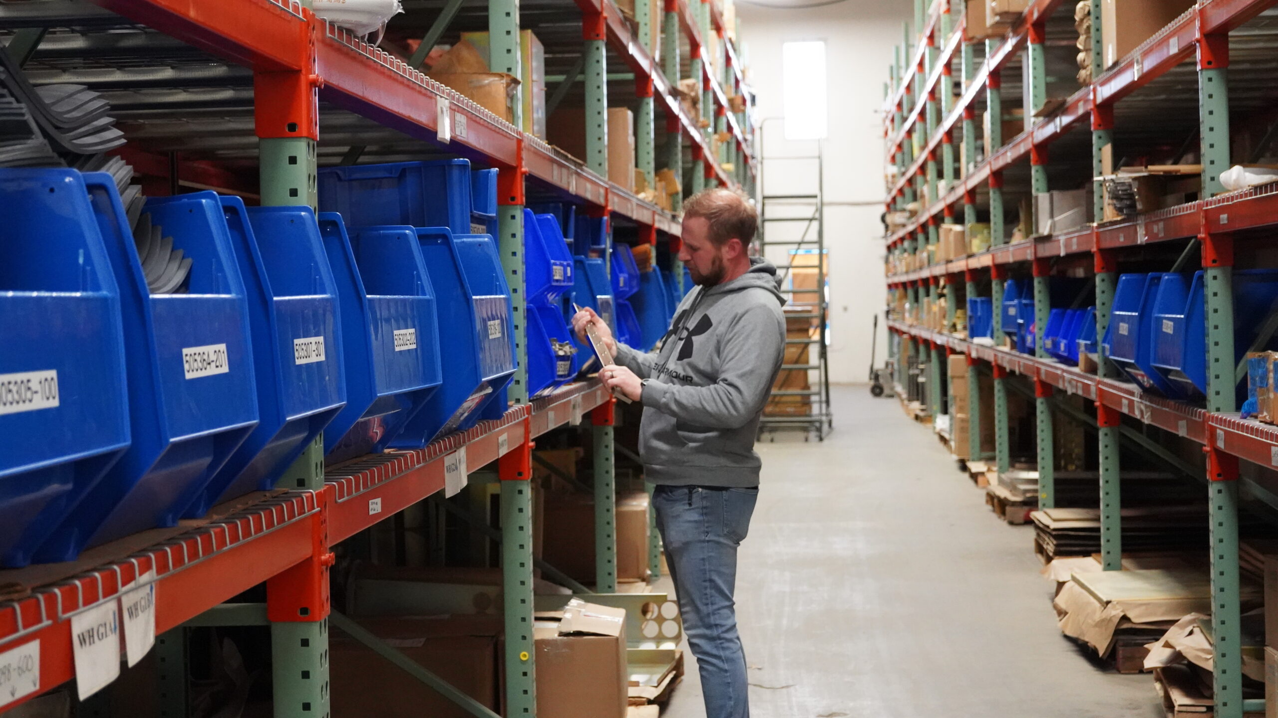 A warehouse worker in a gray hoodie checks a clipboard while selecting items from blue plastic bins on industrial shelving.