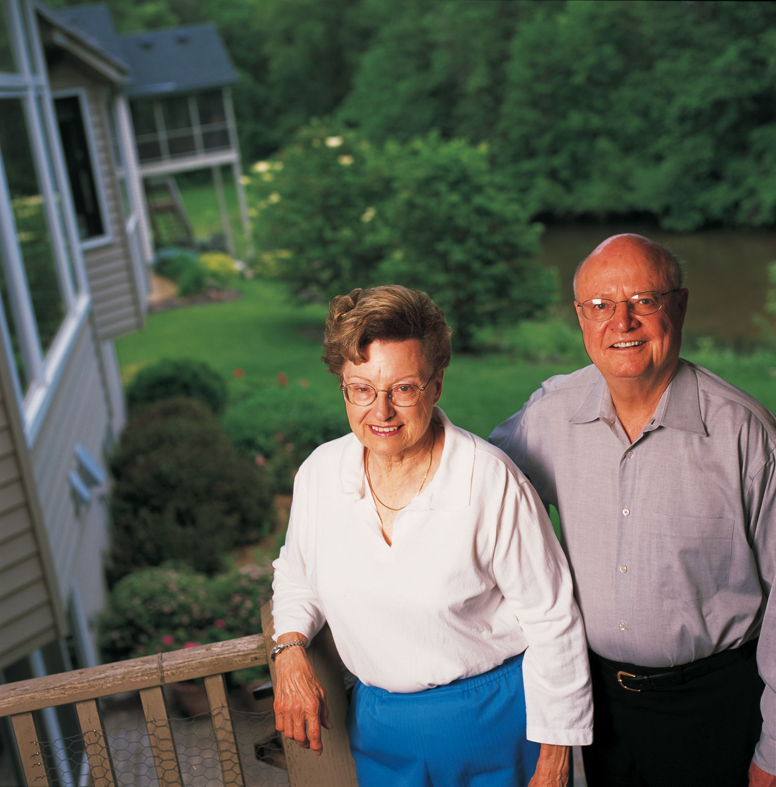 An older couple smiles together while standing on a deck overlooking a yard and wooded area.
