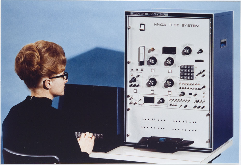 A woman in a black shirt sits at a desk using a keyboard connected to a large Micro Control M-10A Test System console.