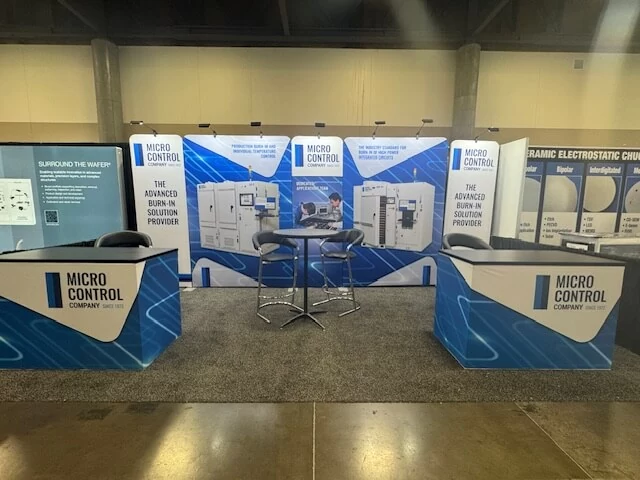 Trade show booth for Micro Control Company featuring a blue and white branded backdrop, two information desks, and a small table with two chairs.