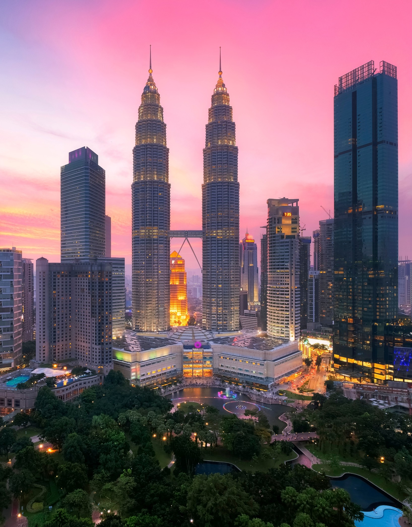 The Petronas Twin Towers illuminated at sunset against a bright pink sky, overlooking a park with fountains and city skyscrapers.