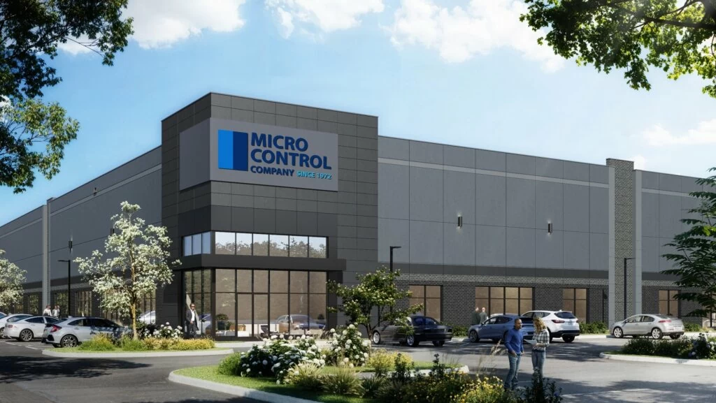 A modern warehouse building featuring the Micro Control Company logo on the exterior, with a parking lot and small groups of people in the foreground.