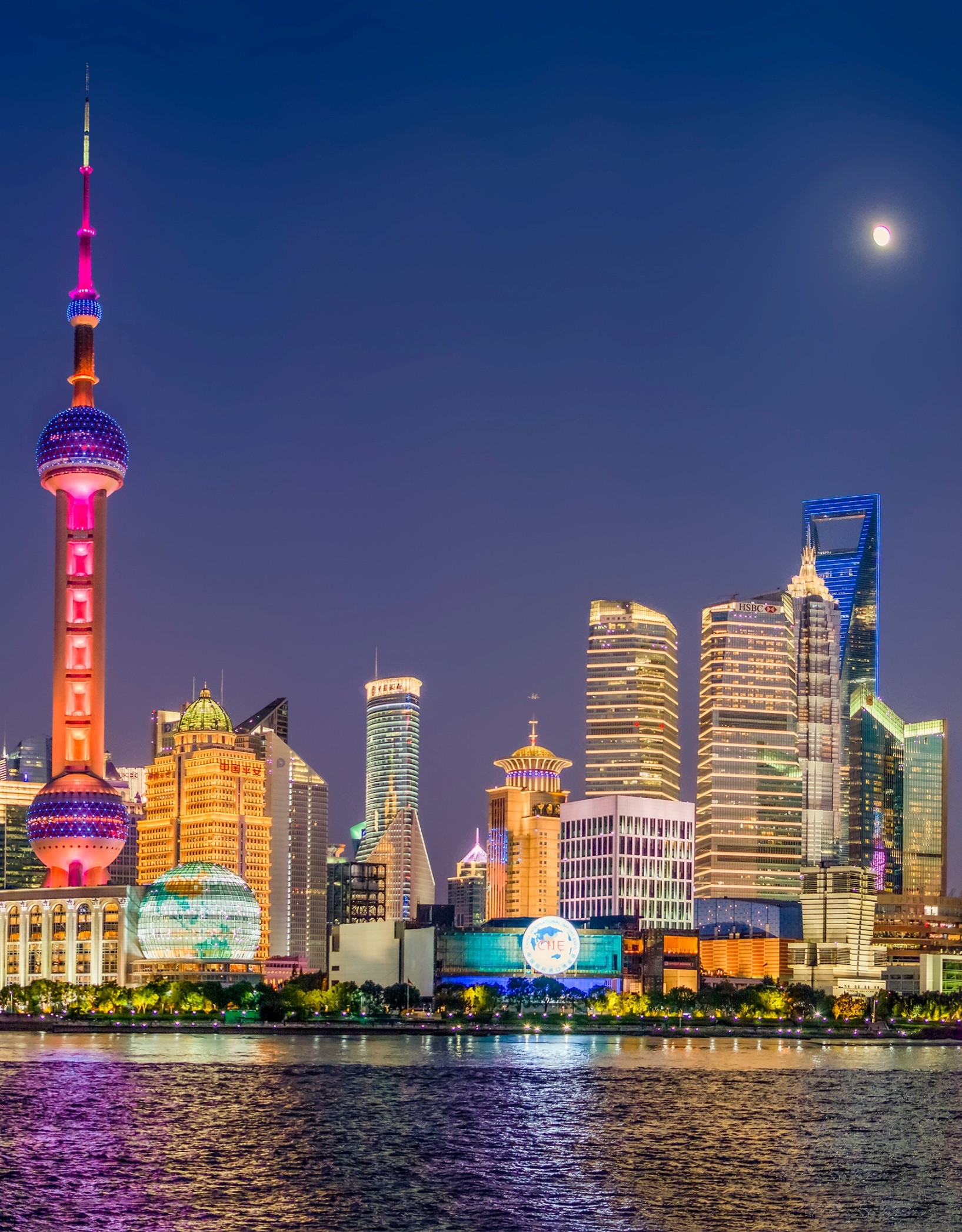 The Shanghai skyline at night featuring the glowing Oriental Pearl Tower and surrounding skyscrapers reflected in the river under a crescent moon.