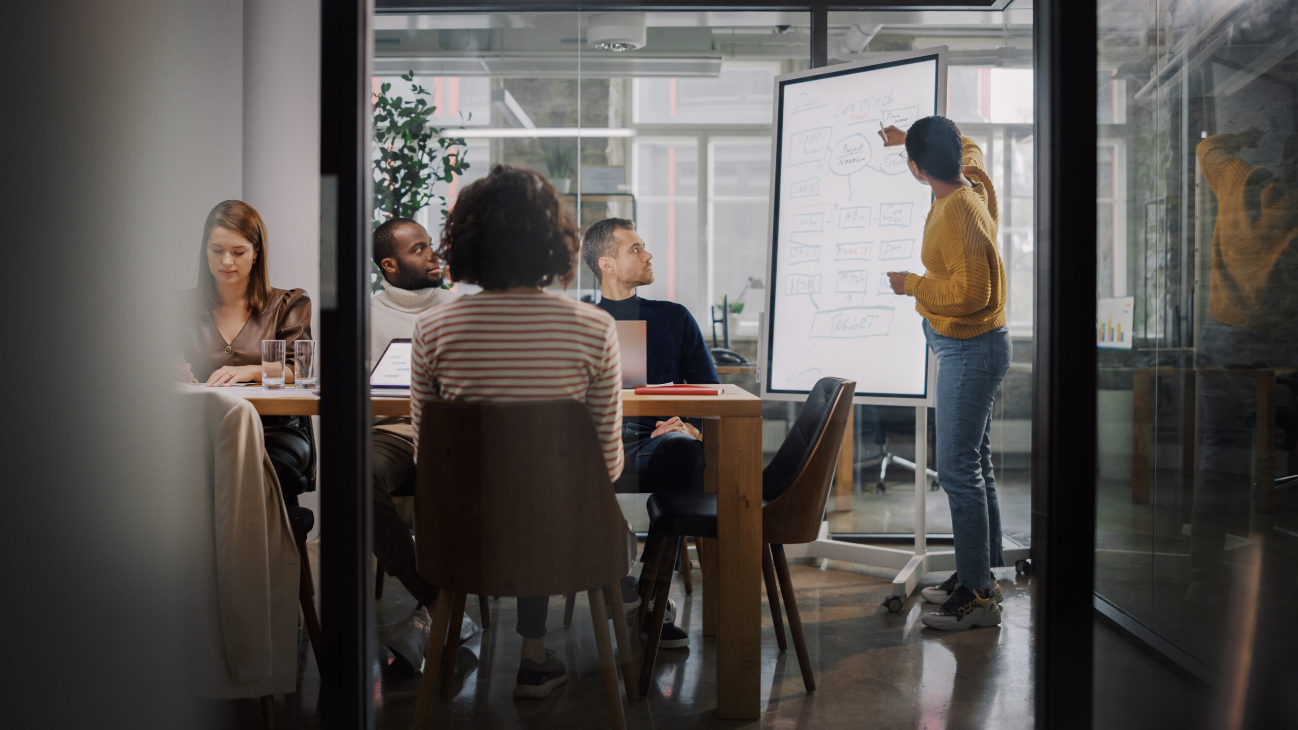 A woman stands at a whiteboard presenting a flowchart to four colleagues seated at a conference table in an office.