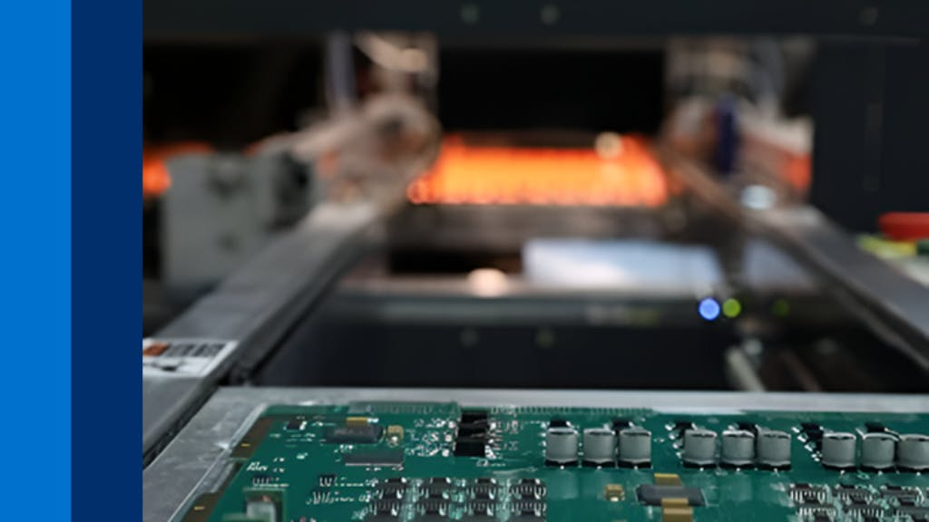 A green circuit board on a conveyor belt moving toward an industrial heating oven.