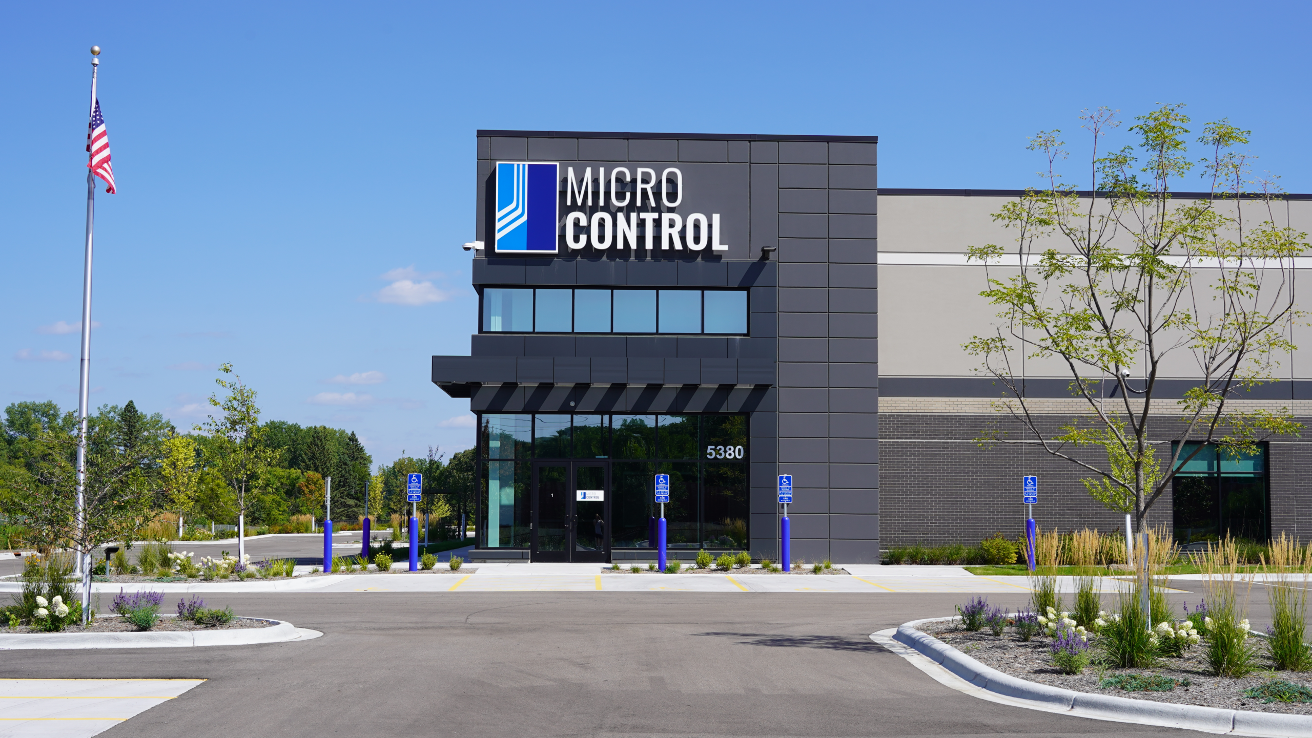 The exterior of the Micro Control office building, featuring a dark facade with the company logo and an American flag flying on a pole in front.