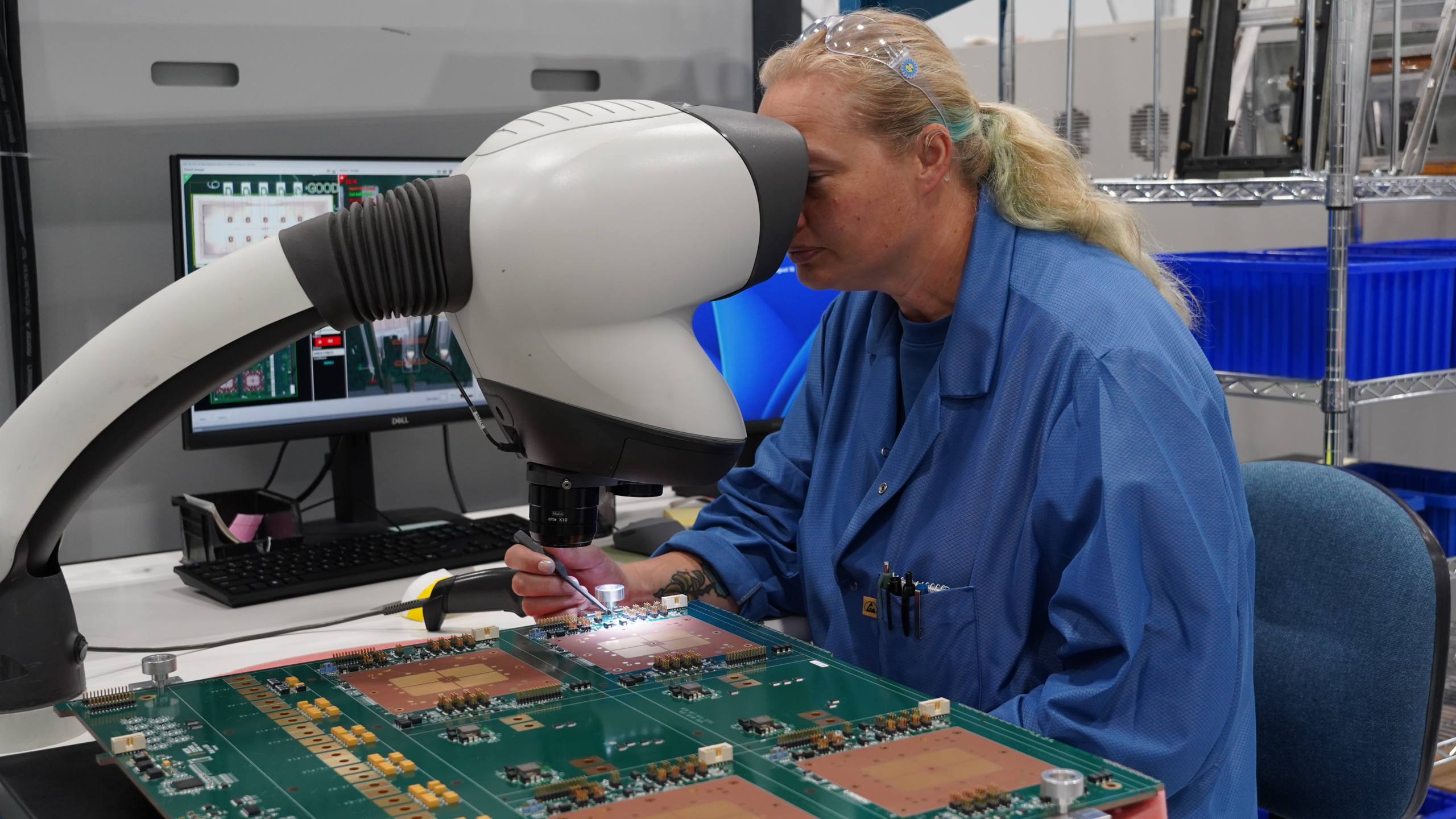 A technician in a blue lab coat uses a digital microscope to inspect a large circuit board.