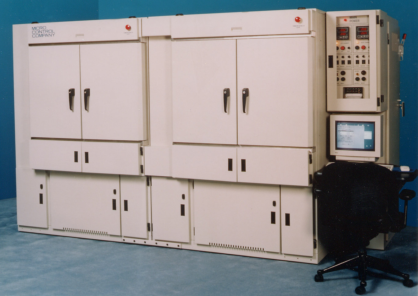Micro Control Company industrial testing system with two large cabinet doors, a control panel with buttons and displays, a computer monitor, and an office chair.