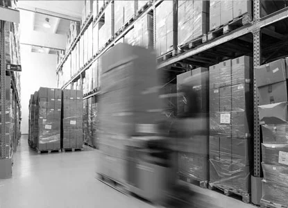 A forklift carrying a tall stack of boxes drives through a warehouse aisle lined with floor-to-ceiling storage racks.